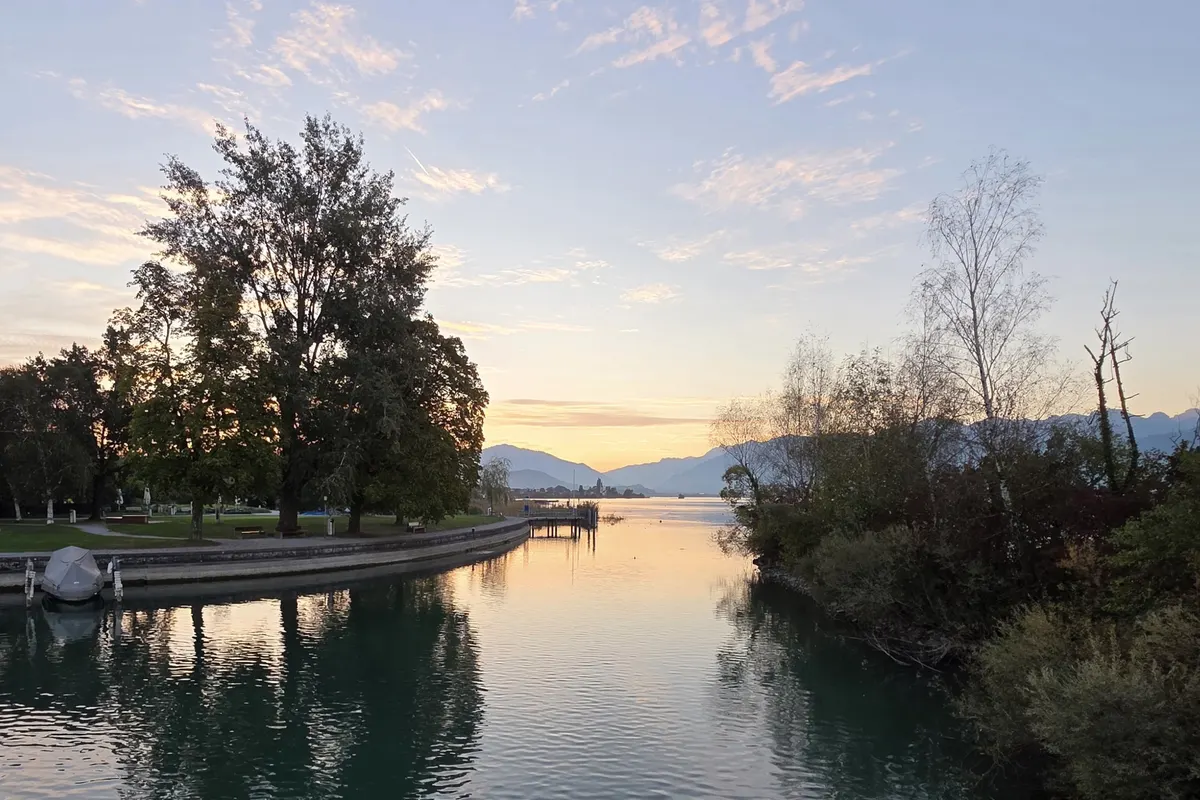 Abendstimmung an einem Seeufer: Bäume spiegeln sich im ruhigen Wasser, dahinter Berge im letzten Licht
