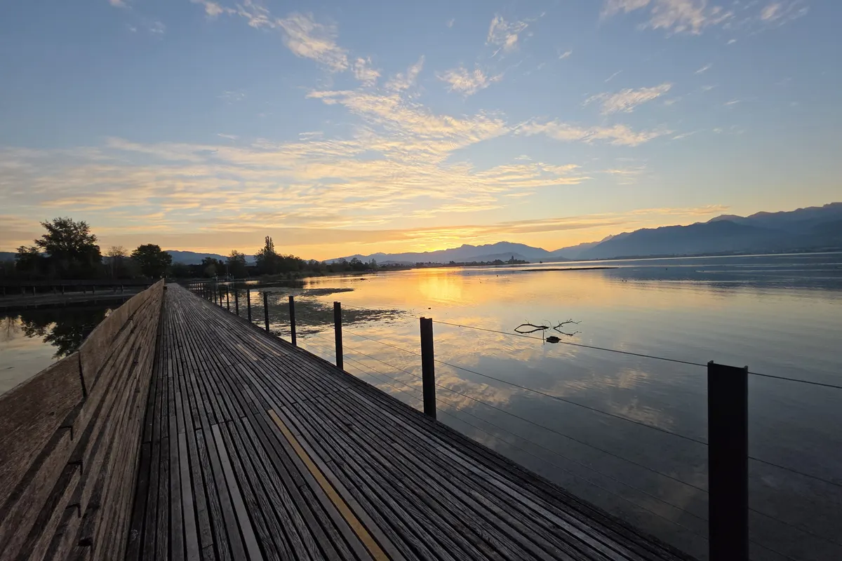 Ein Holzsteg führt bei Sonnenaufgang über den See, das Licht spiegelt sich im Wasser